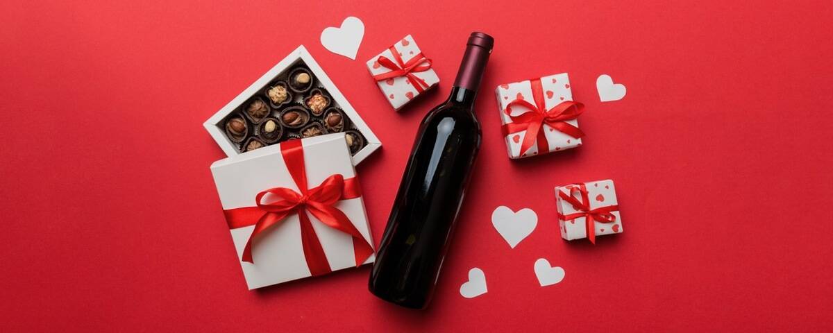 Red wine with chocolates, small gifts, and heart cutouts on a red background, creating a romantic mood.